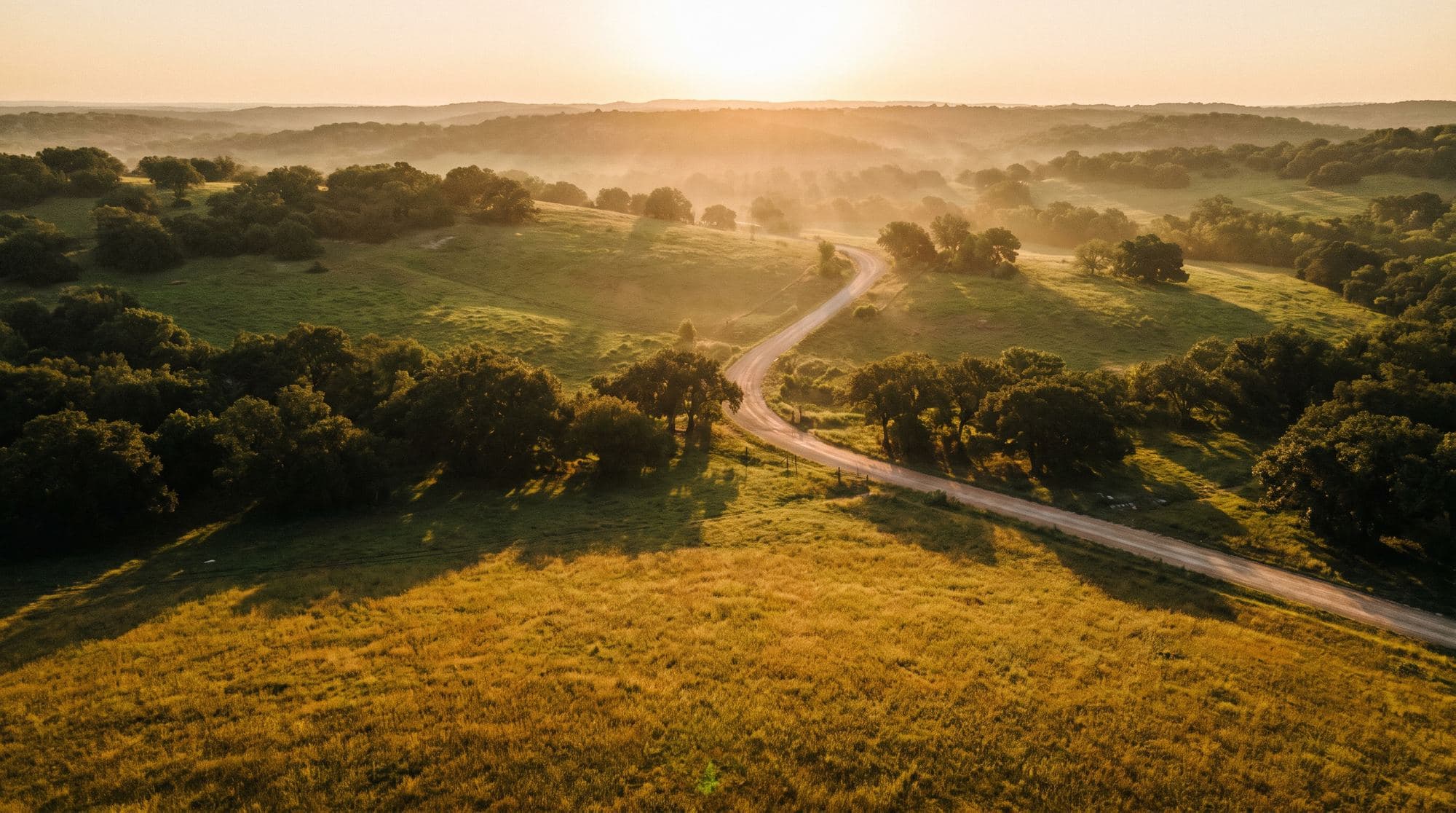 Golden-hour aerial view of Texas Hill Country ranch land with oak trees and a winding caliche road